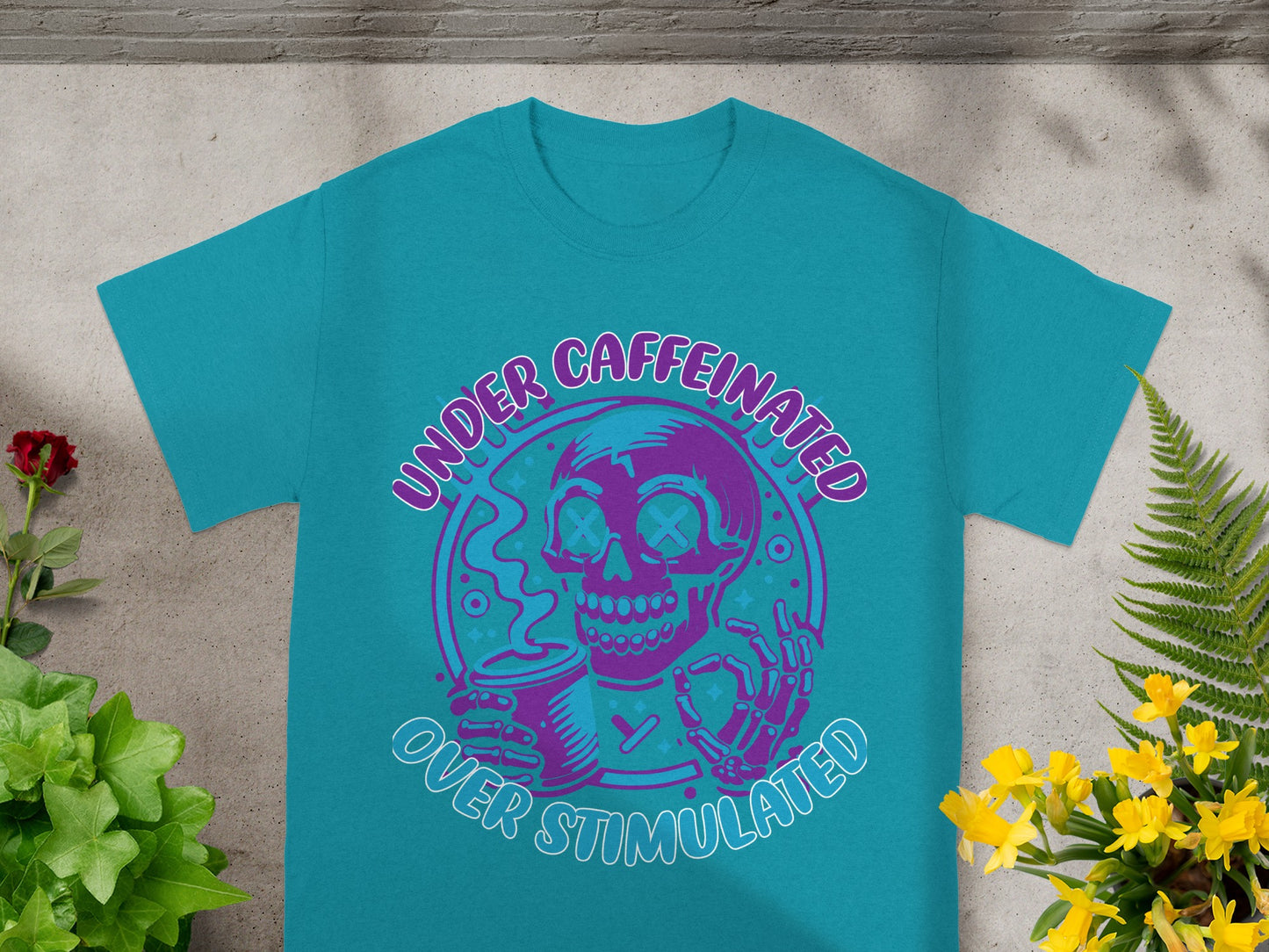 Teal t-shirt with graphic of a skeleton holding coffee cup, text "UNDER CAFFEINATED OVER STIMULATED".