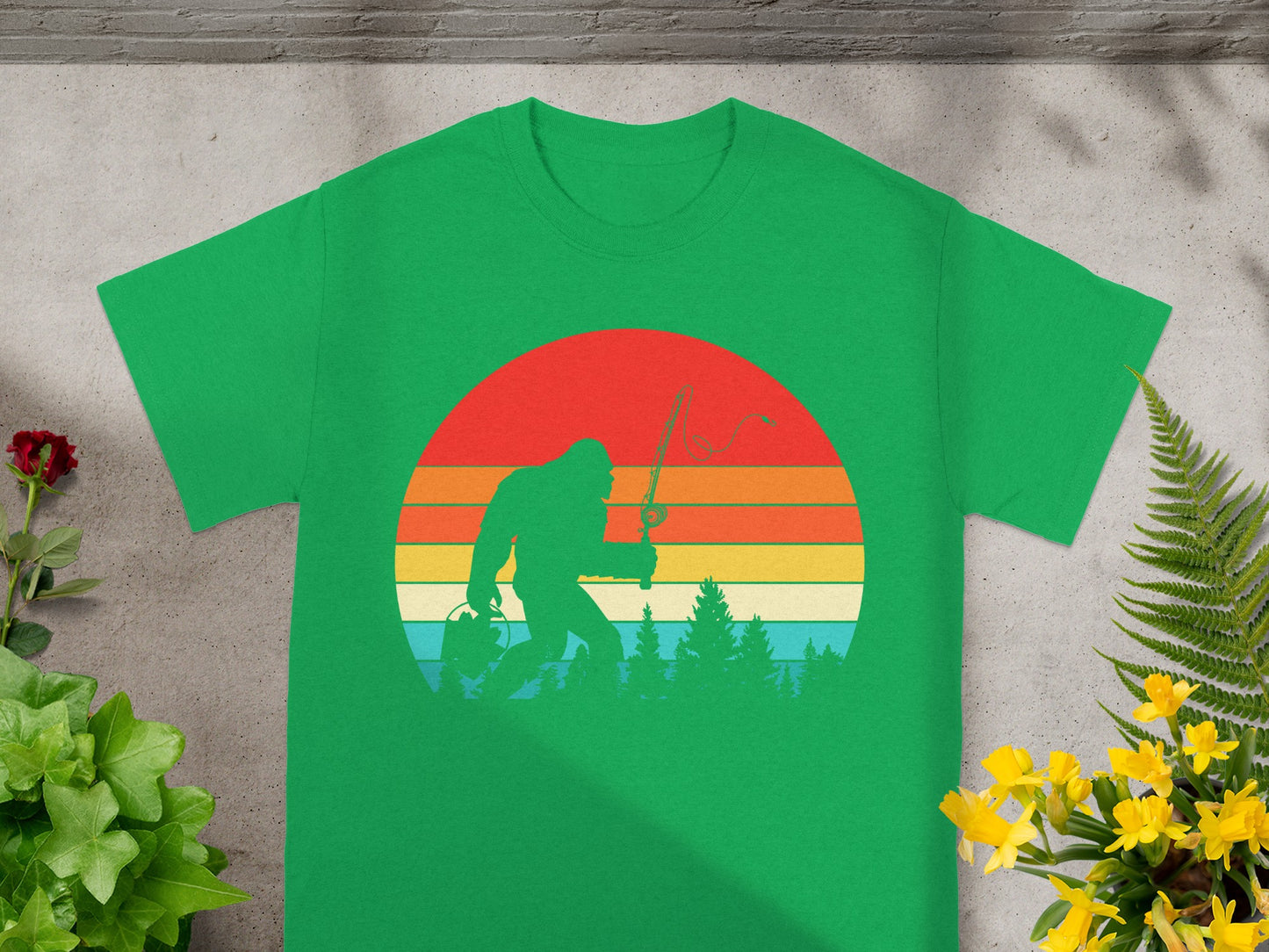 Green t-shirt with Bigfoot fishing graphic against a vintage sunset and forest.