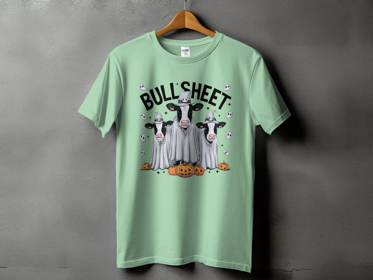 Three cows in ghost costumes with pumpkins and skulls on a light green t-shirt.