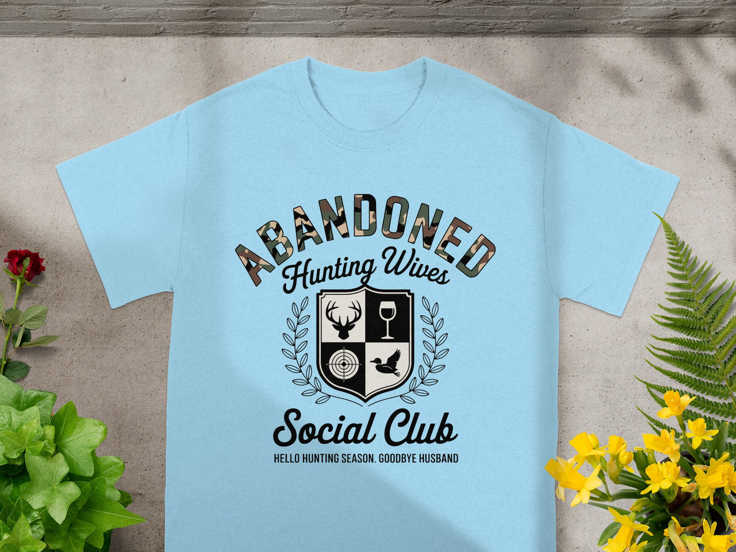 Light blue t-shirt with "Abandoned Hunting Wives Social Club" text and shield graphic.
