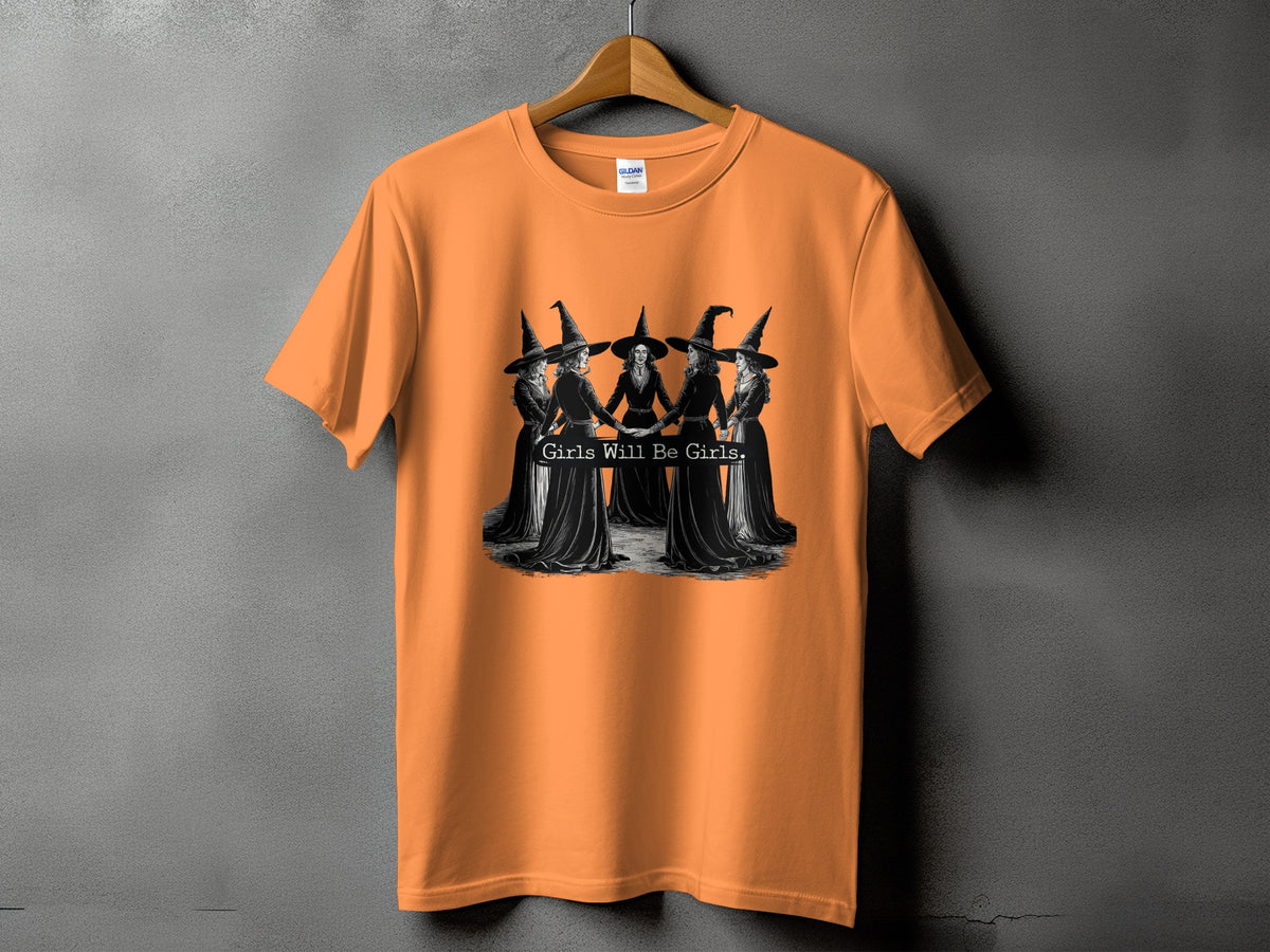 Orange t-shirt with a graphic of witches holding hands and the text "Girls Will Be Girls."