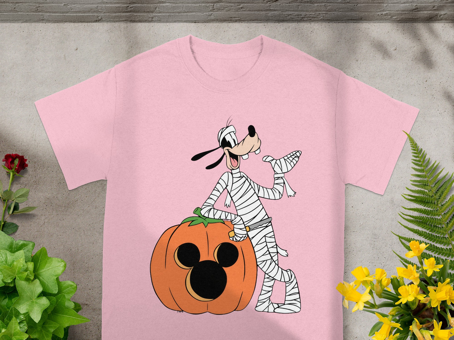 Goofy dressed as a mummy leaning on a Mickey Mouse pumpkin on a pink t-shirt.