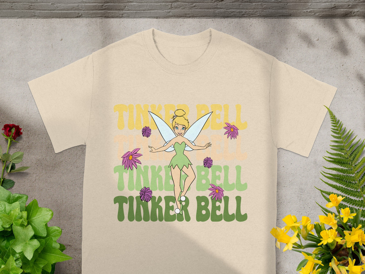 Tinker Bell illustration with bold text "TINKER BELL" on a light beige t-shirt.