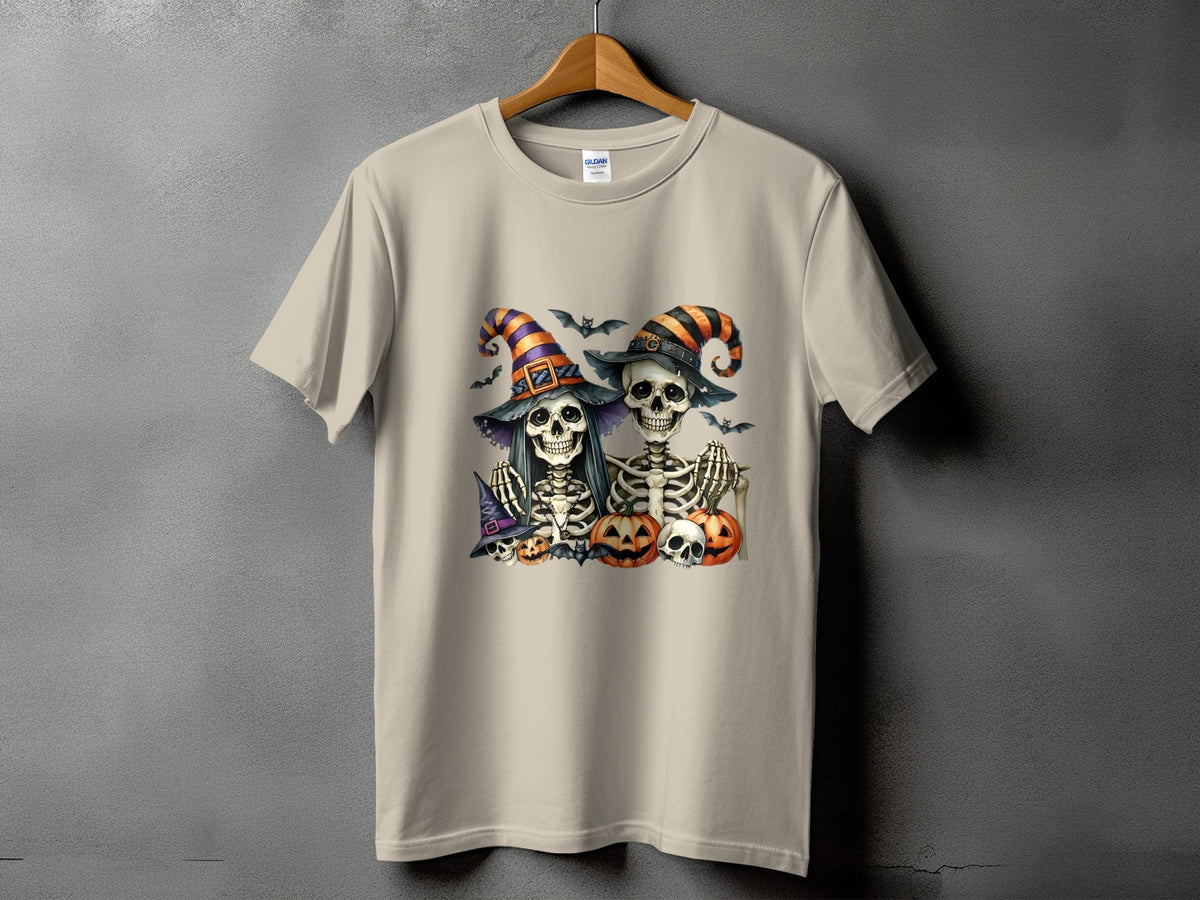 Two skeletons in witch hats with pumpkins and bats on a tan t-shirt.