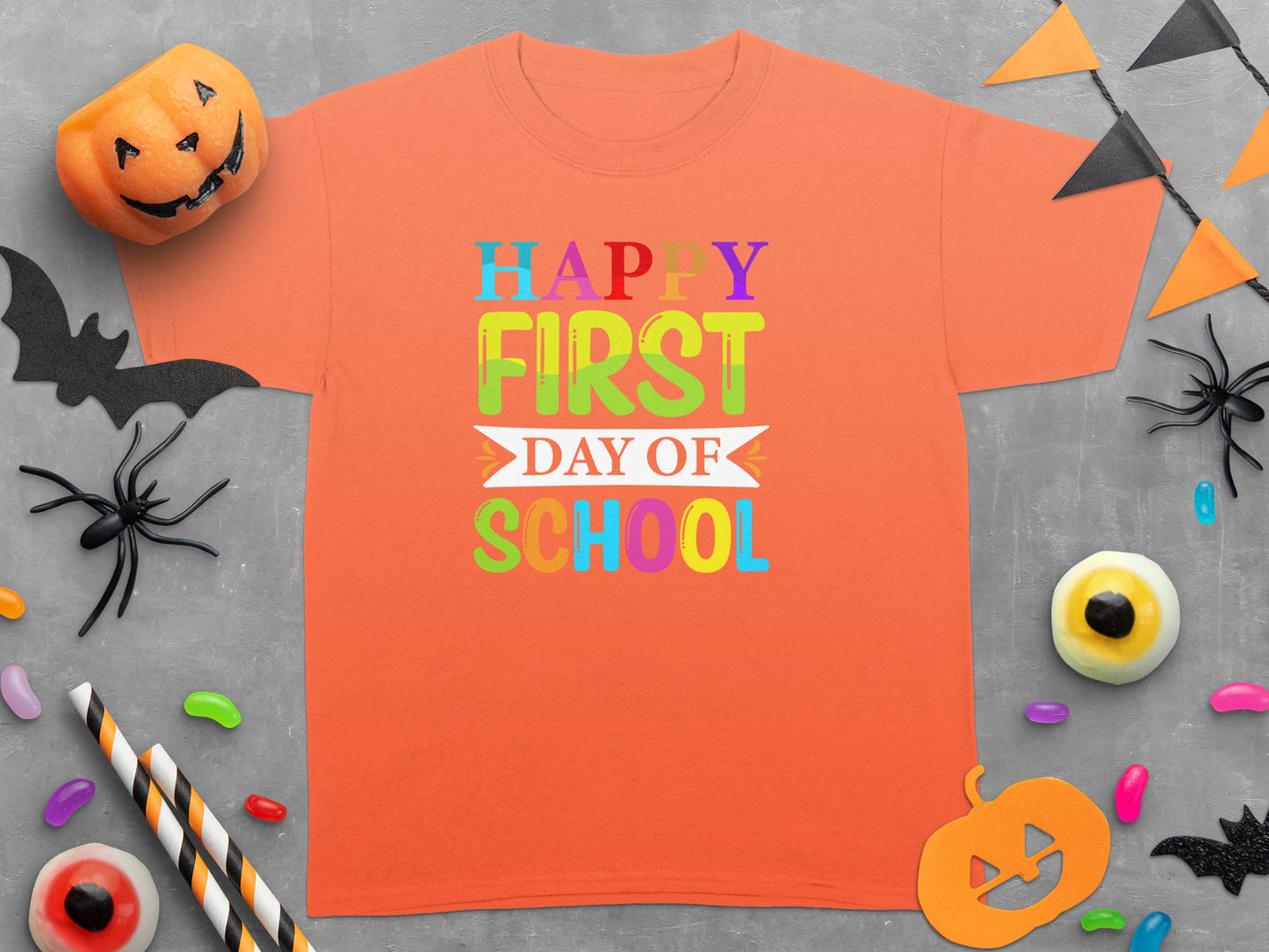 Orange t-shirt with colorful "Happy First Day of School" text, surrounded by Halloween decorations.