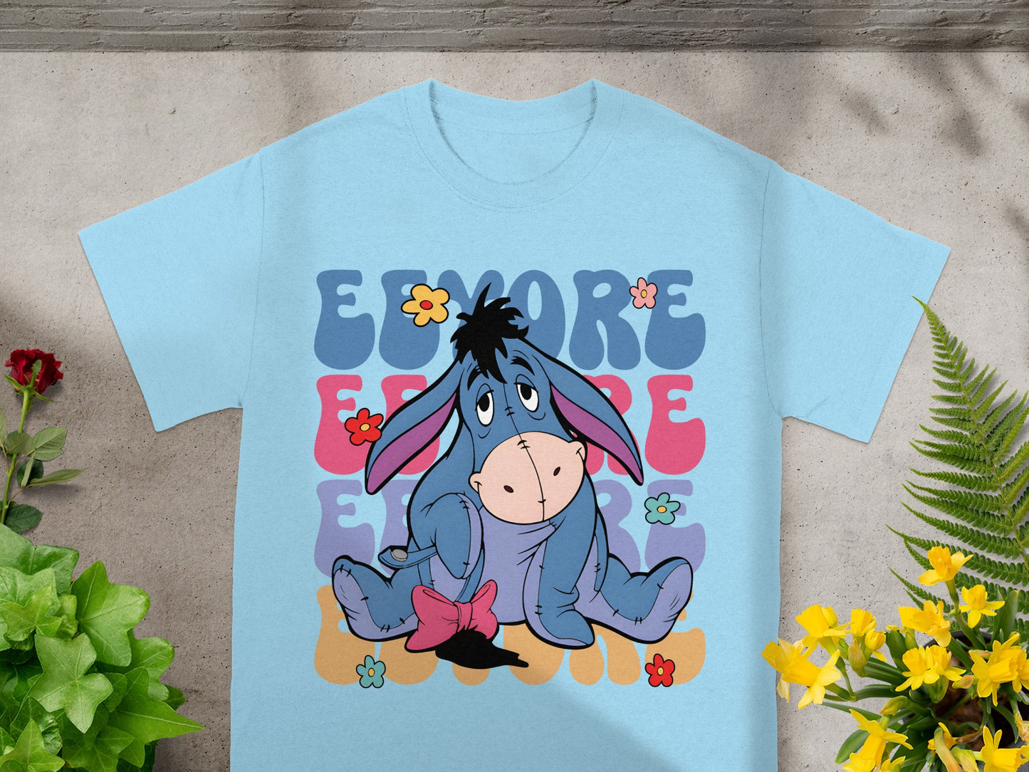 Eeyore sitting among colorful flowers and text on a light blue t-shirt.