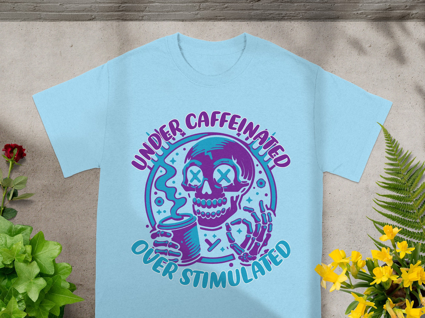 Light blue t-shirt with skeleton holding coffee and text "UNDER CAFFEINATED OVER STIMULATED".