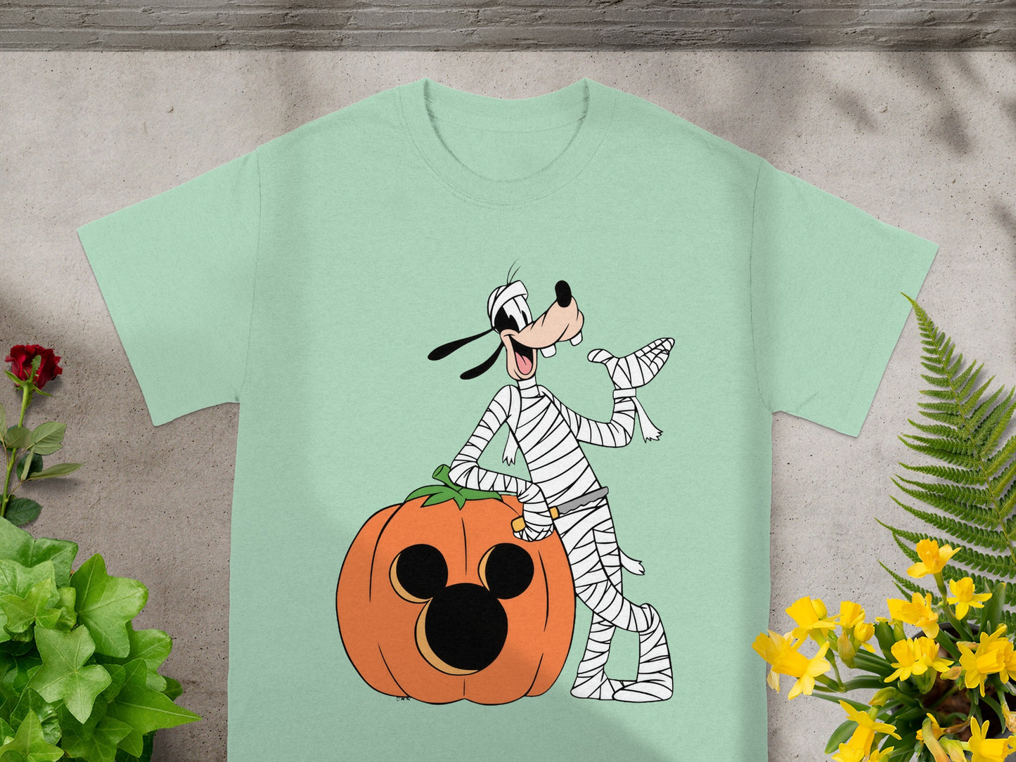 Goofy in mummy costume leaning on a Mickey Mouse pumpkin on a light green t-shirt.